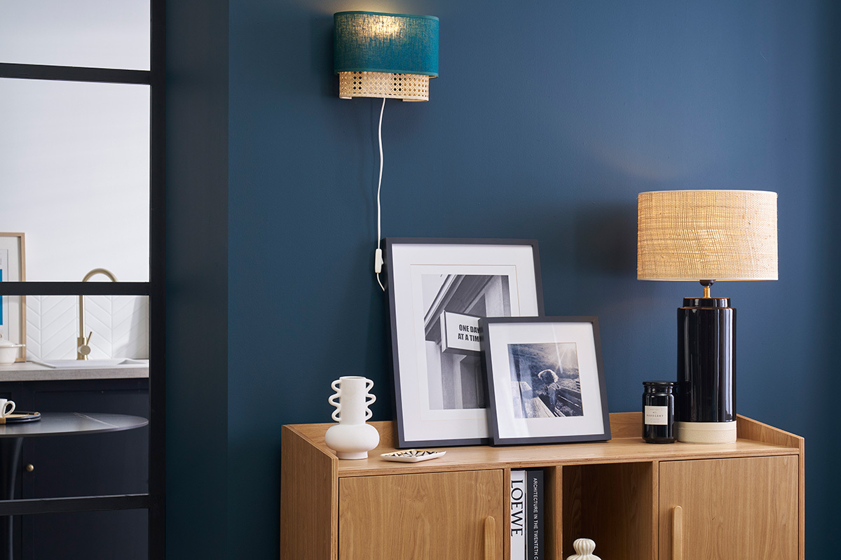 Applique murale bleu canard et rotin sur mur bleu, � c�t� de cadres et lampe sur meuble.
