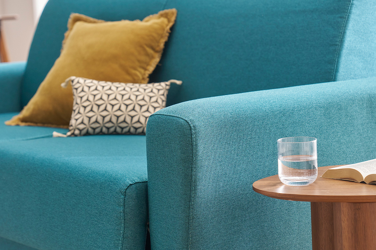Canap� convertible bleu canard avec coussins, vue de c�t�, table en bois avec verre et livre.