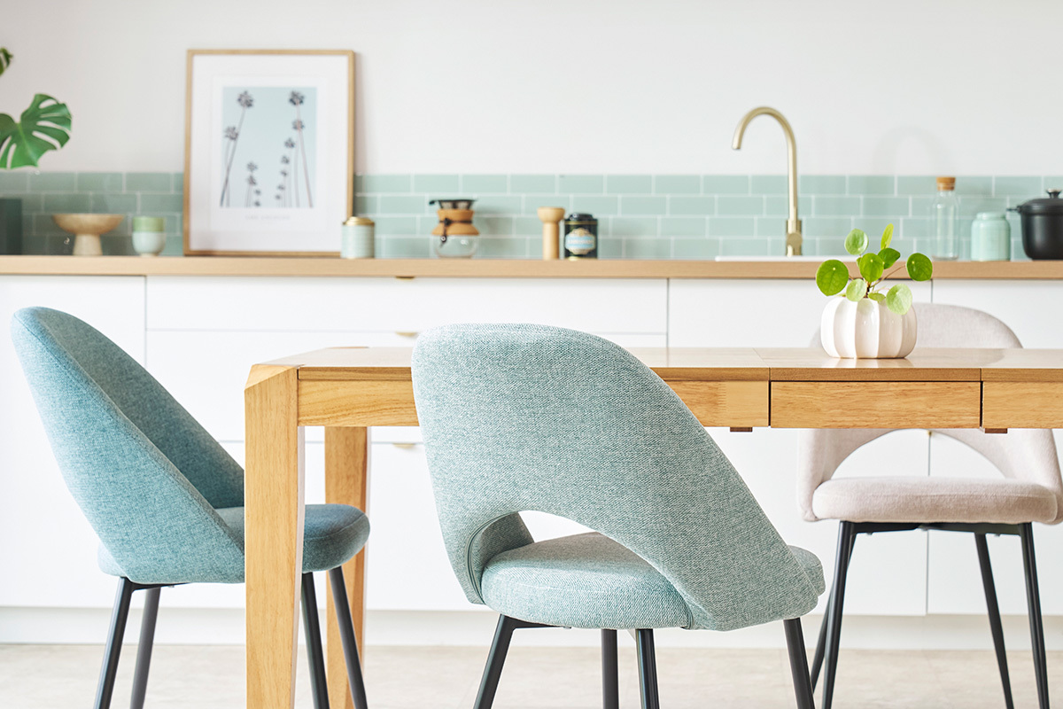 Chaises en tissu vert c�ladon avec pieds noirs, vue de dos, autour d'une table en bois clair dans une cuisine moderne.