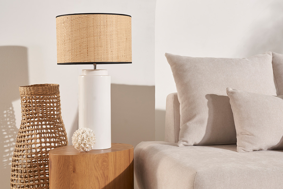 Lampe � poser blanche en c�ramique avec abat-jour en raphia sur table, � c�t� d'un canap� et d'une d�coration en rotin.