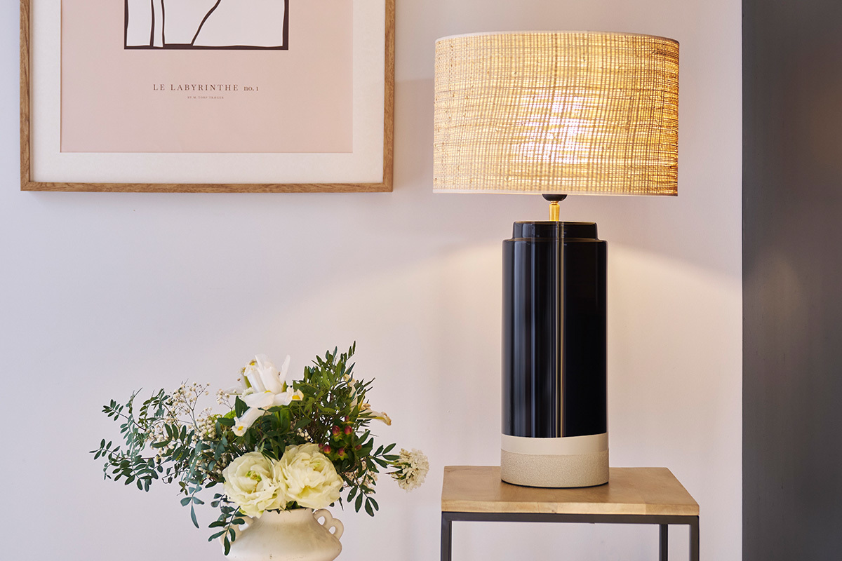 Lampe � poser noire en c�ramique avec abat-jour en rabane, pos�e sur une table � c�t� de fleurs.