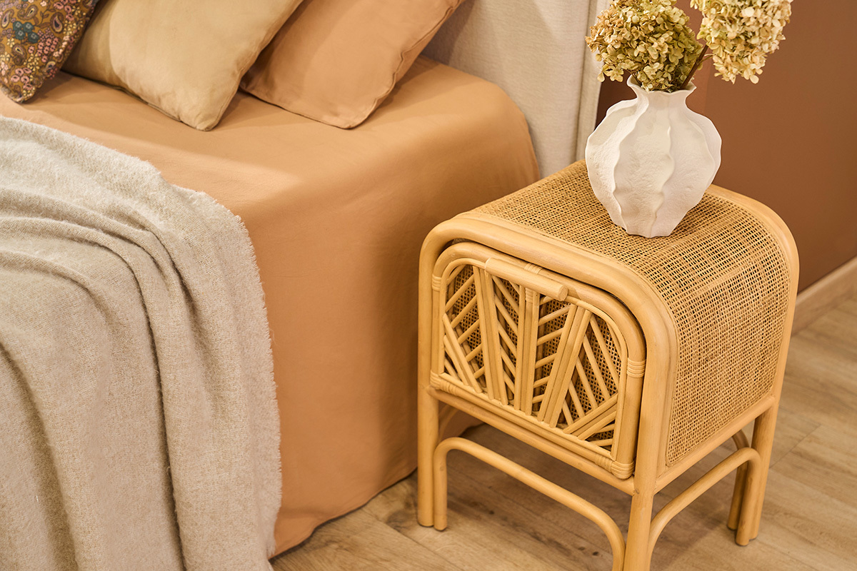 Table de chevet en rotin GALON avec motif, vue lat�rale, � c�t� d'un lit avec vase blanc.